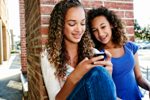 Two teenage girls lean against a brick wall tooking at a cell phone and smiling.