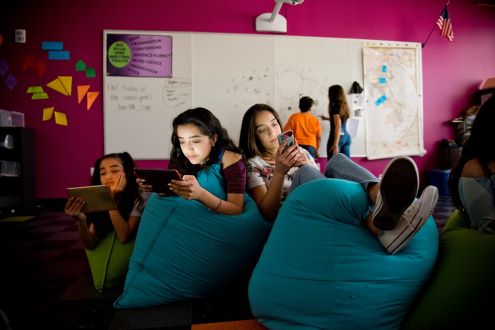 three students sit on soft chairs while looking at devices