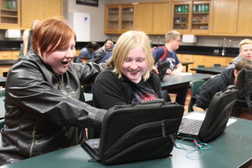 Dos niños en un aula se cuestionan mutuamente en un portátil.