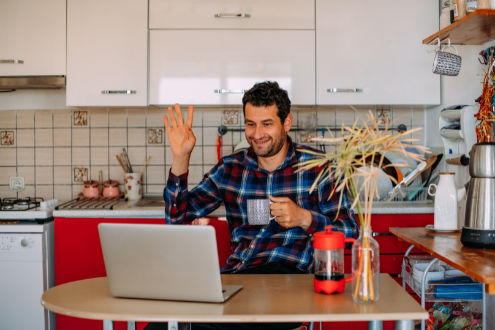 Un hombre se sienta en una computadora portátil en su cocina saludando a las personas que asisten a una conferencia virtual.