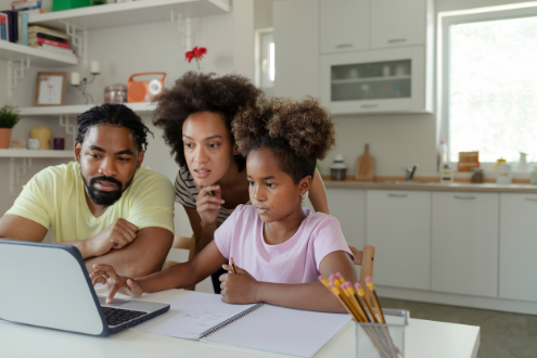 Two parents help a daughter with homework on a laptop in the kitchen