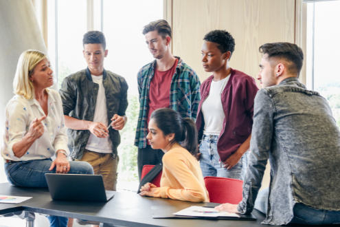a teacher sits on a desk surrounded by five students who she's speaking to