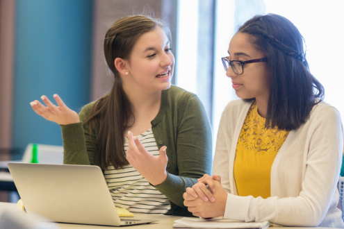 Two girls at school in front of a laptop discuss cyber-bullying incident.