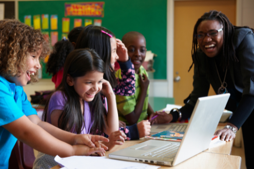 A group of children enthusiastically look at a laptop while their laughing teacher looks on. 