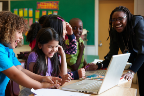 A group of children enthusiastically look at a laptop while their laughing teacher looks on.