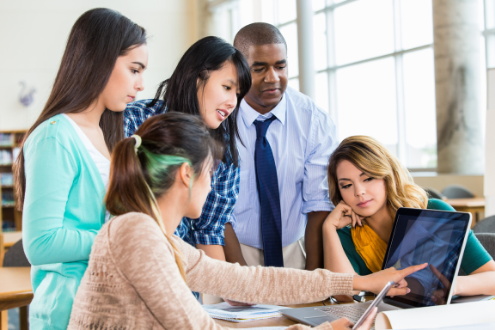 A teacher looks on as four female students discuss a school project around a computer screen
