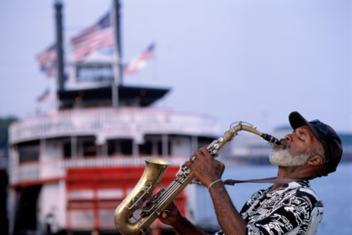 A man plays saxaphone with a riverboat in the background in New Orleans
