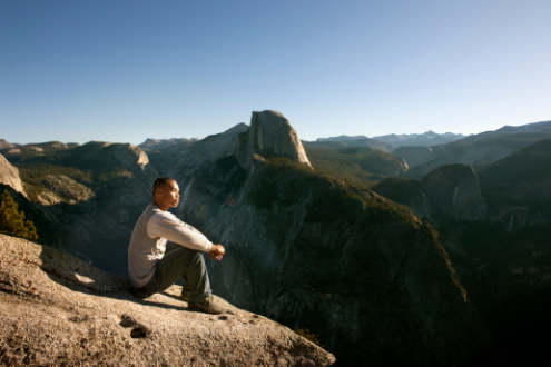 A man sits on the top of a cliff looking out into a gorgeous valley