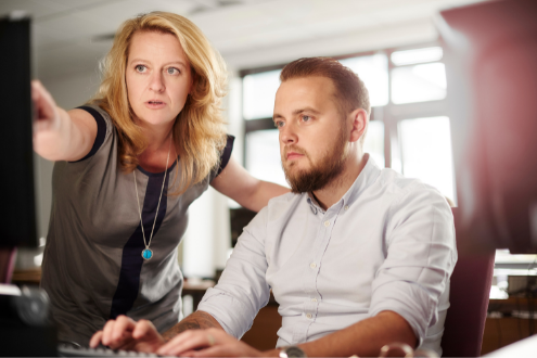 A woman standing points to the screen of a desktop computer while a man sits and observes.