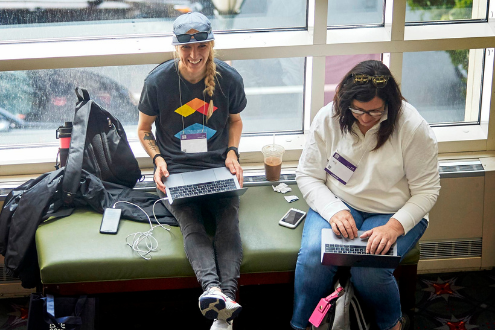 Two women sit on a window sill seat smiling while working on laptops at ISTE19.