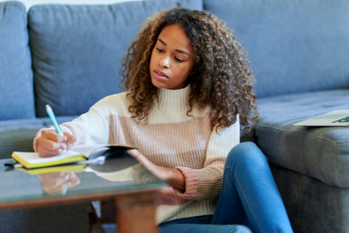 a woman sits on the floor of her home and writes in a notebook while holding papers