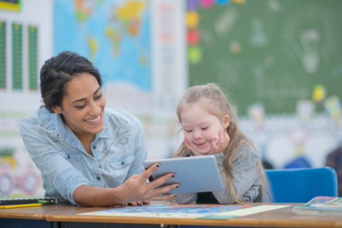 A teacher helps a young girl with Down Syndrome use an iPad in a classroom