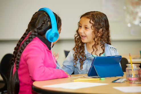 Two girls facing each other sitting at a desk in a classroom with an iPad. One girl is wearing headphones.