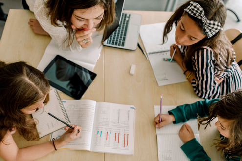 A group of students sit together at a table with papers and tablets working on a school project