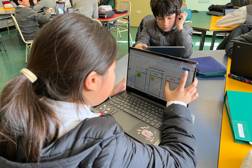 una niña y un niño en un salón de clases sentados en escritorios mirando sus computadoras portátiles de la escuela