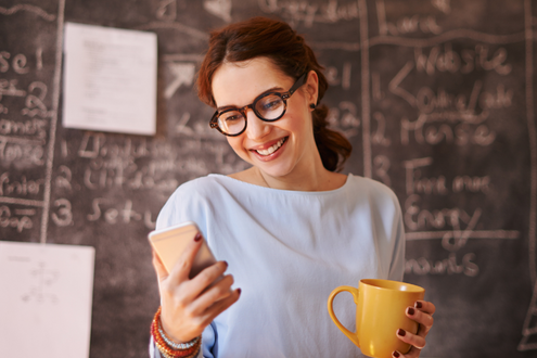 una maestra mira su teléfono sonriendo mientras sostiene una taza de café