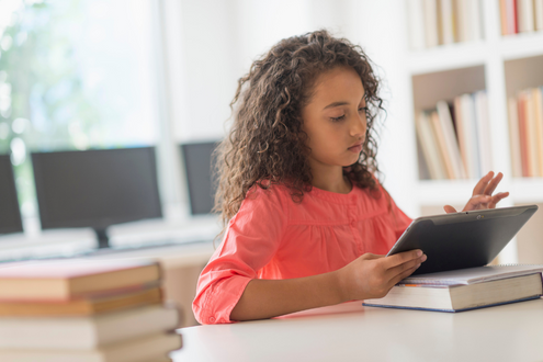 a girl sits at a table in a classroom doing work on a computer tablet