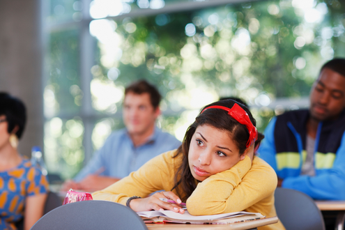 A girl sits at a school desk with her head resting on her arm looking bored