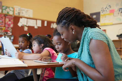 A teacher sits at a desk next to a young student in a classroom to help her with an assignment on an iPad