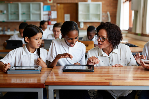 two students at a desk looking down at an iPad while their teacher looks on