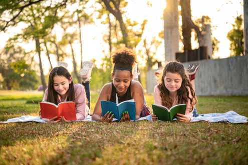 Tres niñas tiradas en el césped afuera leyendo libros