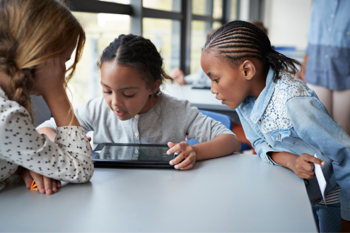 Tres niñas sentadas alrededor de una mesa en un salón de clases mirando un iPad