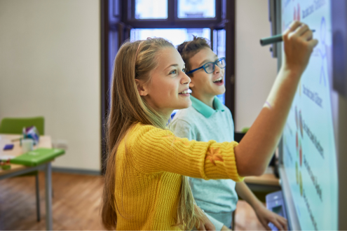 Dos estudiantes se ríen mientras escriben algo en una pizarra digital en un salón de clases.