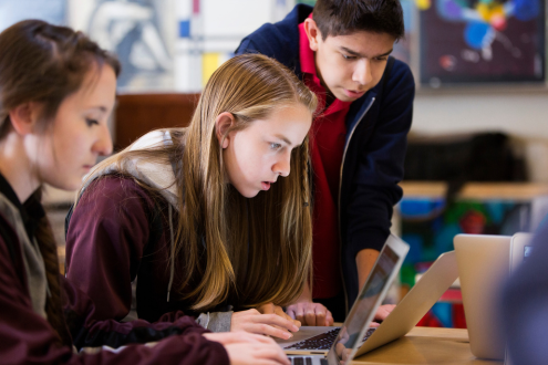 Three students sit at a desk in a classroom looking intently at a laptop.