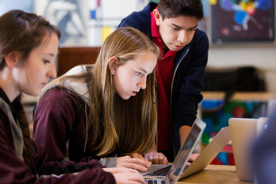 Tres estudiantes se sientan en un escritorio en un salón de clases mirando fijamente una computadora portátil.