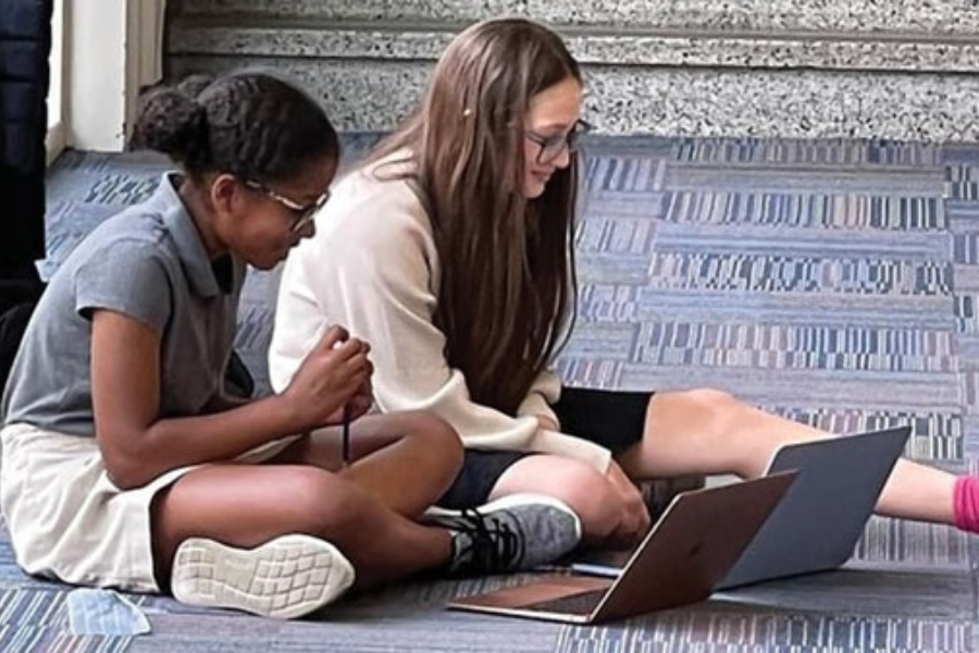 Two girls sit side by side on the floor of a classroom working on their laptops