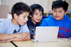 Three boys in a classroom sit side by side watching something on a laptop. 